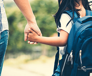 Kind mit Rucksack läuft Hand in Hand mit einer erwachesenen Person.