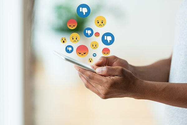 Person hält Handy in den Händen über dem traurige und wütende Emojis zu sehen sind. 