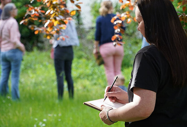 Frau im Vordergrund macht sich Notizen. Drei Personen stehen im Hintergrund im Garten.