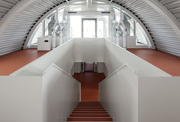 Zu sehen ist der helle Dachraum mit sichtbarer Stahlkonstruktion des Tonnendachs sowie der Treppenaufgang in weißem Anstrich und rotbraunem Linoleum-Bodenbelag.