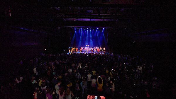 Foto vom Finale des Musikwettbewerbs 'Ecole du Flow' während der Europawochen, aufgenommen im Tollhaus. Das Bild zeigt eine belebte Szene mit vielen Zuschauern, die einer Performance auf einer beleuchteten Bühne in einer dunklen Halle beiwohnen.