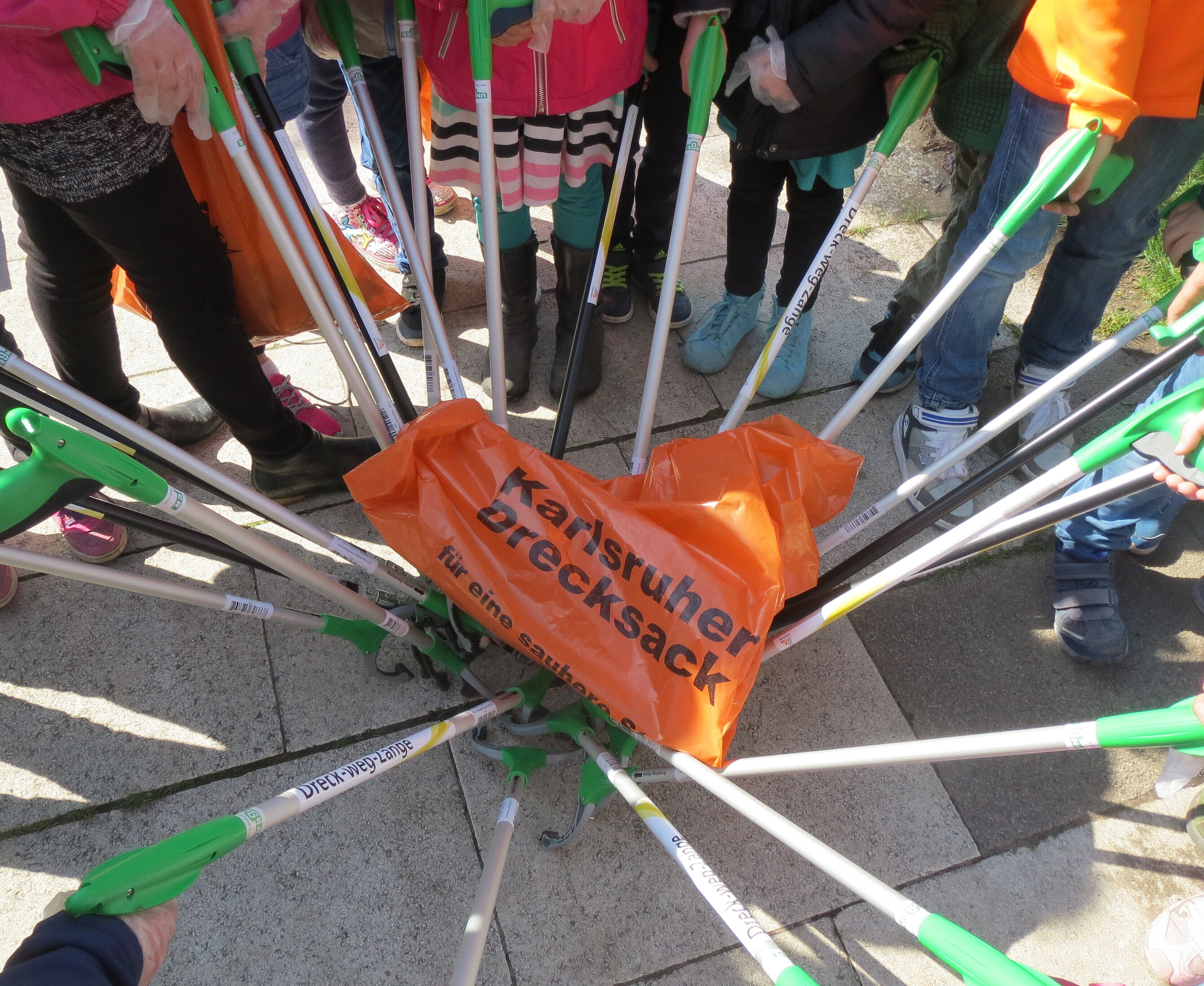 Dreck-Weg-Wochen Helferinnen und Helfer halten ihre Abfallzangen in alle in die Mitte. Darauf liegt ein "Drecksack" der Stadt Karlsruhe.