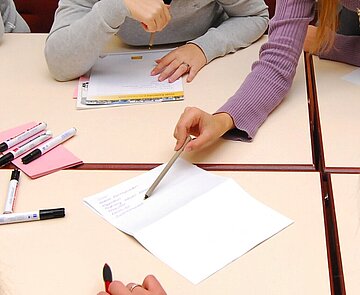Zusammenarbeit Person zeigt am Tisch mit Schreibgerät auf ein Schriftstück.