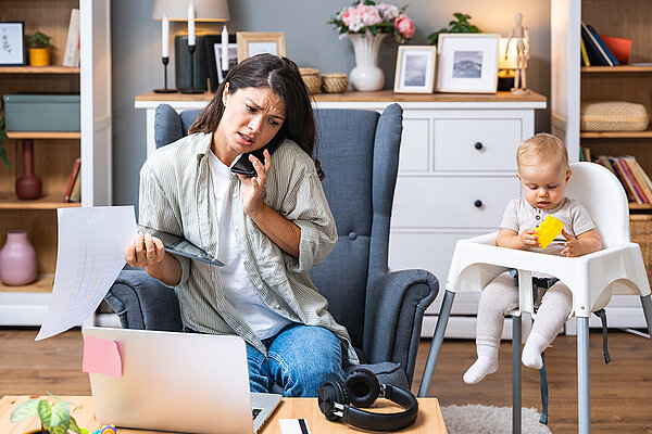 Junge gestresste Mutter kümmert sich um ihr kleines Baby und arbeitet von zu Hause aus am Laptop. 
