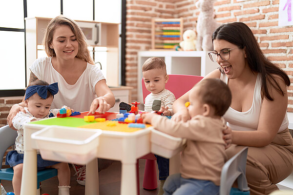 Zwei junge Frauen sitzen mit drei Kindern am Tisch und spielen mit Bausteinen