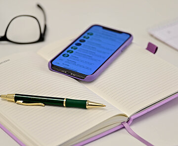 Terminvereinbarung Bild zeigt einen Tisch. Auf diesem liegen ein Notizbuch, ein Smartphone, ein Kuli, eine Brille. Im Smartphone-Display ist ein Kalender zu sehen.