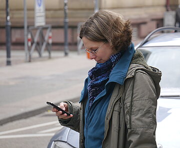 KA-Feedback Das Bild zeigt eine Frau, die auf der Straße neben einem parkenden Auto steht, mit einem Smartphone in der Hand.
