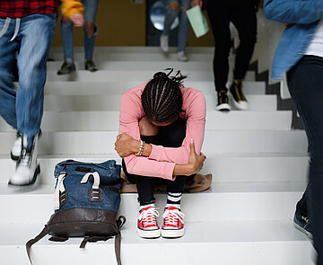 Prävention an Schulen Jugendliche Person sitzt mit angezogenen Beinen und Kopf auf den Knien auf der Schultreppe, während Mitschülerinnen und Mitschüler vorbeilaufen.