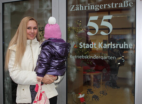 Junge Frau mit Kind auf dem Arm steht vor dem Kindergarten der Stadt Karlsruhe