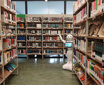 Karlotta, der Roboter der Bibliothek,  steht zwischen vollen Bücherregalen