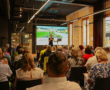 Bild von den Teilnehmenden Konferenz in bestuhlten Reihen, vorne auf der Bühne spricht Bürgermeisterin Bettina Lisbach