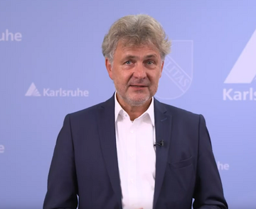 Oberbürgermeister Dr. Frank Mentrup vor blauem Hintergrund mit Fächerwappen der Stadt.