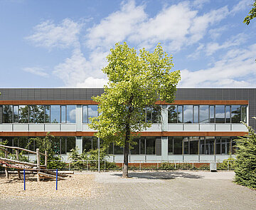 Das Bild zeigt den Schulhof und die anthrazitfarbene Südfassade, die mittig von einem kleinen Baum bedeckt ist.