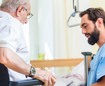 Pflegekraft hilft älterem Mann aus dem Bett in den Rollstuhl.