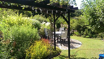 Das Bild zeigt eine Sitzgelegenheit zwischen Stauden und Rasen mit einer Pergola in der Kleingartenanlage SChloss.