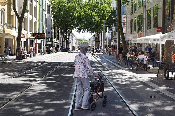 Das Bild steht symbolisch für Menschen mit eingeschränkter Mobilität. Es zeigt eine ältere Dame mit Gehhilfe, die eine Straße in der Karlsruher Innenstadt überquert.