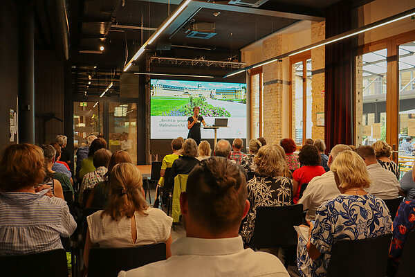 Bild von den Teilnehmenden Konferenz in bestuhlten Reihen, vorne auf der Bühne spricht Bürgermeisterin Bettina Lisbach