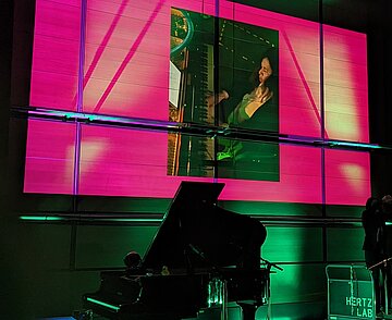 Dunkle Bühne mit zwei Flügeln und einer Pianistin, die über eine Leinwand zu sehen ist