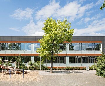Das Bild zeigt den Schulhof und die anthrazitfarbene Südfassade, die mittig von einem kleinen Baum bedeckt ist.