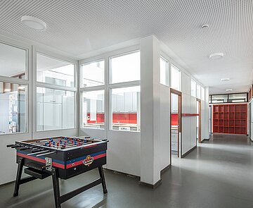Zu sehen ist der helle, moderne Flurbereich mit Kautschukboden und abgehängter Gipskarton-Lochdecke, der teilweise als Spielbereich mit Tischkicker genutzt wird.