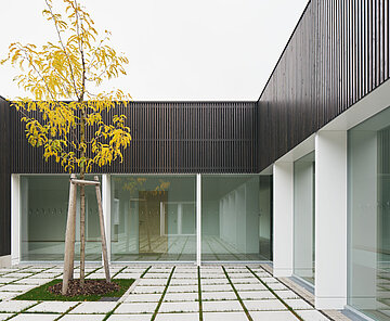 Blick vom Innenhof auf das Rasenpflaster und einen frisch gepflanzten Baum, dahinter ein Teil der dunkelgrauen Holzlattenfassade mit großen Fenstern und weißen Rahmen.