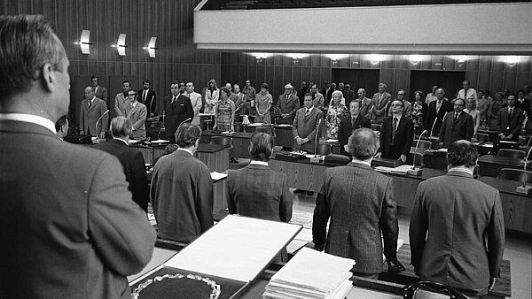 Blick in den Bürgersaal des Rathauses mit den während der Gedenkminute stehenden Fraktionsmitgliedern. Im Vordergrund die Regierungsbank mit den stehenden Mitgliedern.