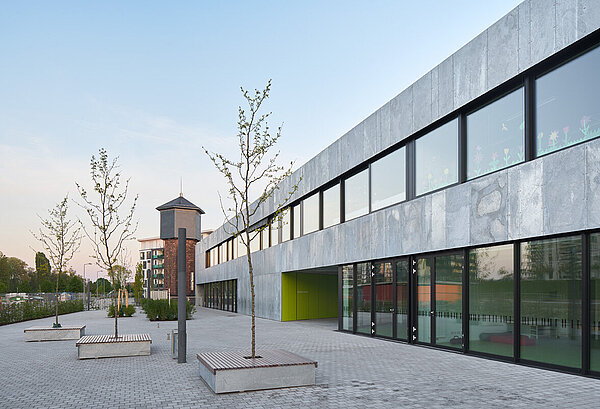 Zu sehen ist die helle Nordfassade mit großen Fensterflächen, Sitzgelegenheiten und im Hintergrund der Wasserturm.