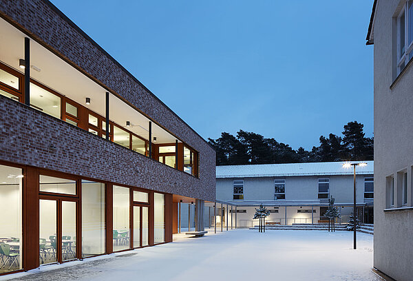 Zu sehen ist die beleuchtete Südansicht des Schülerhorts mit rotbraun-weißem Sichtziegelmauerwerk, Fenstern und Türen in hellbronze sowie dem überdachten Weg, der zum Altbau führt.
