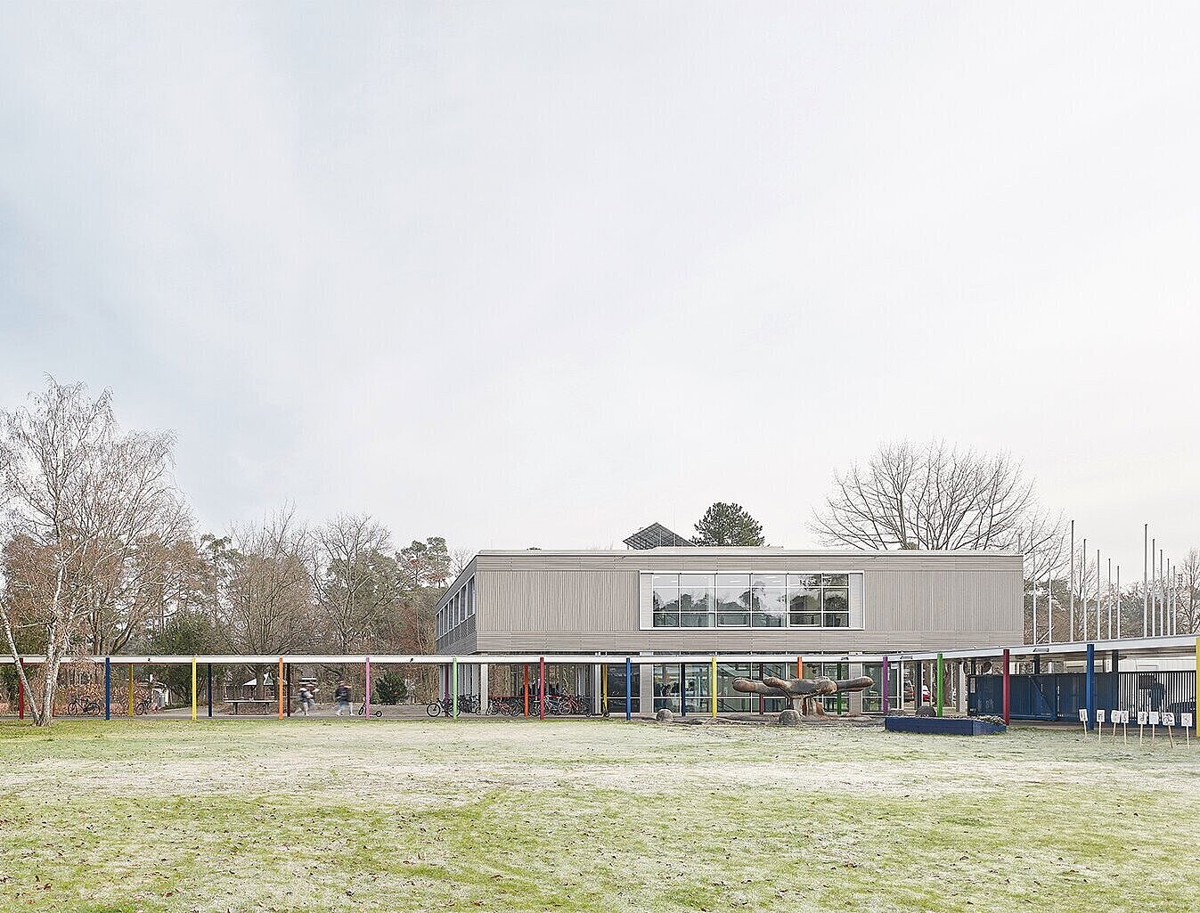 Zu sehen ist die Westansicht des Gebäudes D in grauen Tönen sowie ein überdachter Weg für die Schüler, der von bunten Stützen getragen wird.