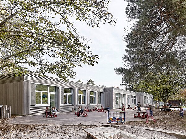 Das Bild zeigt die Südperspektive der Schule Gebäude A mit der grau lasierten Holzfassade und spielenden Kindern im Spielgarten.