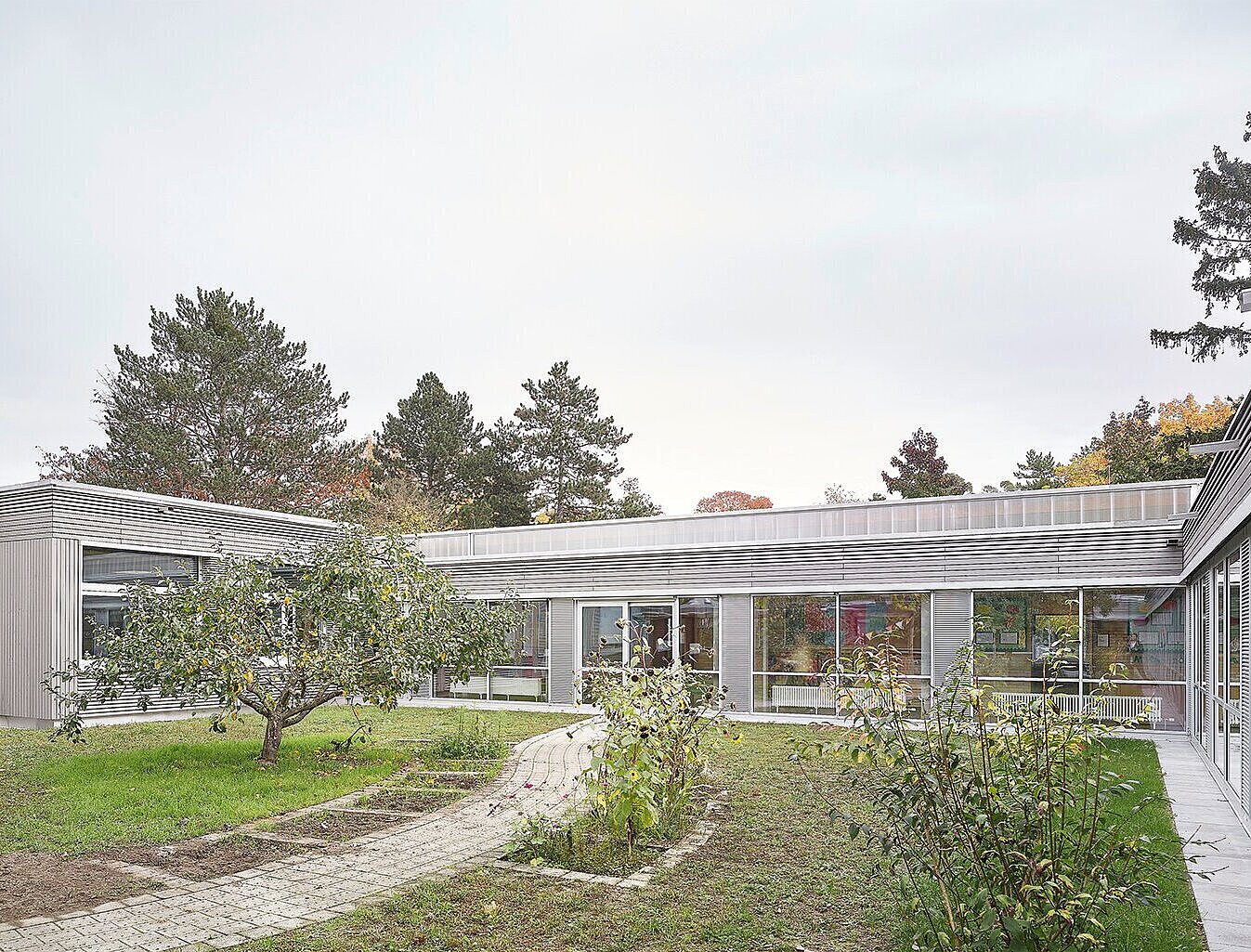 Das Bild zeigt die grau lasierte Nordfassade aus Holz mit großen Fensterflächen und den Schulgarten.