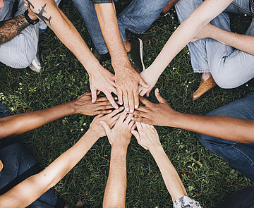 Das Bild zeigt Jugendliche auf einer Wiese, sie sitzen im Kreis und strecken die Hände in der Mitte zusammen.