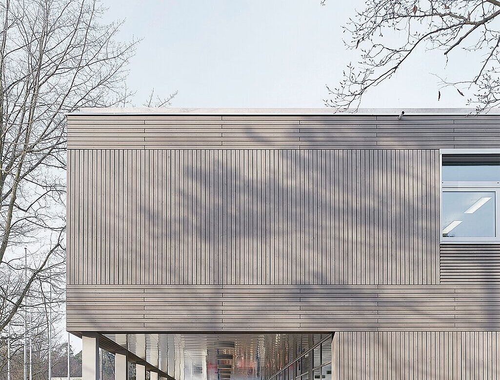 Das Bild zeigt eine Detailansicht der grau lasierten Holzfassade und ein überdachter Weg für die Schüler, der durch das darüberliegende, vorspringende Geschoss entsteht.
