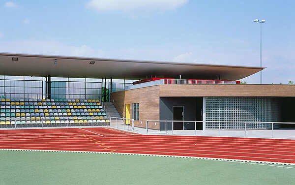 Zu sehen ist die überdachte Tribüne sowie der angrenzende Funktionsbau, dessen Fassade aus Douglasienholzbrettern und Glasbausteinen besteht.