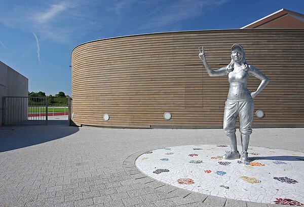 Blick auf die lebensgroße Statue „Victoria“ sowie einen Teil des Funktionsbaus, dessen Fassade aus Douglasienholzbrettern besteht.