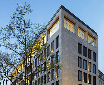 Zu sehen ist die Südostperspektive bei Dämmerung, mit einer Fassade aus Glas und Kalkstein.