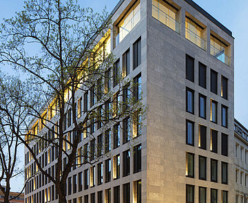 Zu sehen ist die Südostperspektive bei Dämmerung, mit einer Fassade aus Glas und Kalkstein.