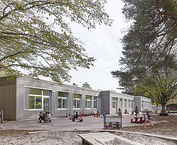 Das Bild zeigt die Südperspektive der Schule Gebäude A mit der grau lasierten Holzfassade und spielenden Kindern im Spielgarten.