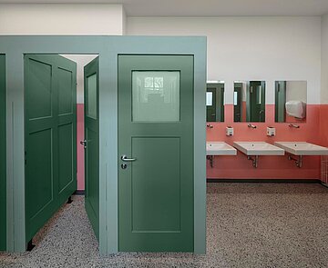 Im Vordergrund sind die sanierten WC-Türen in dunkelgrün abgebildet. Rechts davon hängen mittig an der Wand, weiße Waschbecken mit übergeordneten Spiegeln. Die Wand ist teilweise in weiß und rose gestrichen. Der neu überarbeitete Terrazzoboden vervollständigt die Gestaltung des Raumes. 