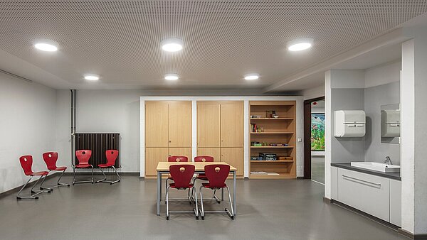 Das Bild zeigt den hellen Gruppenraum mit neuem Kautschukboden, Gipskarton-Lochdecke mit energieeffizienten LED-Leuchten und sanierter Waschnische. Der Raum bietet viel Platz für eine individuelle Möblierung.