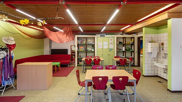 Zu sehen ist ein alter Gruppenraum vor der Sanierung mit Holzdecke und Linoleumboden. Der Raum ist mit verschiedenen Sitzmöglichkeiten eingerichtet und verfügt zusätzlich über eine Waschnische.