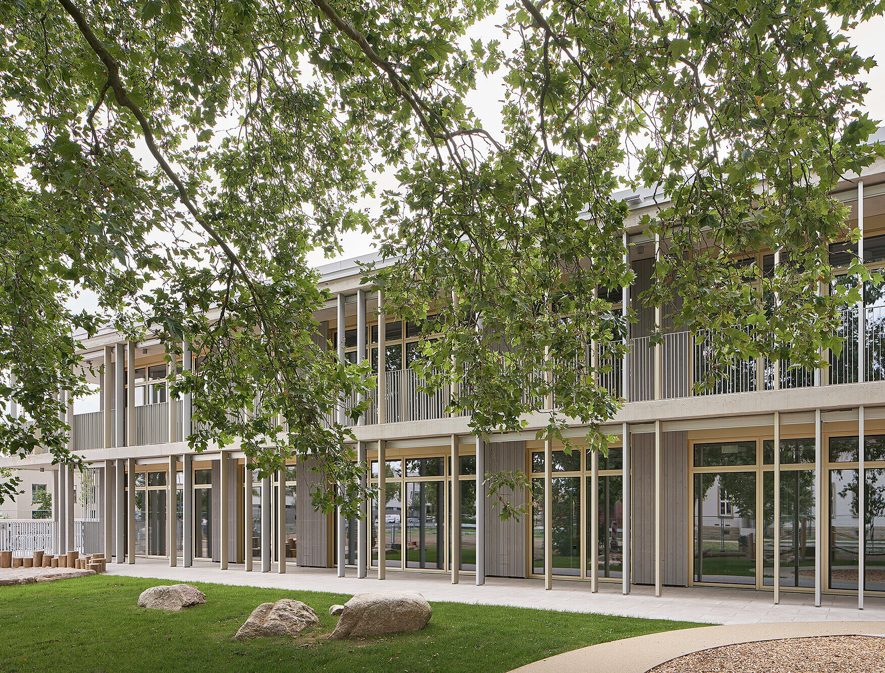 Zu sehen ist ein Teil der Südostfassade mit heller Holzverkleidung und großen Fenstern, die teilweise von einem Baum verdeckt wird.