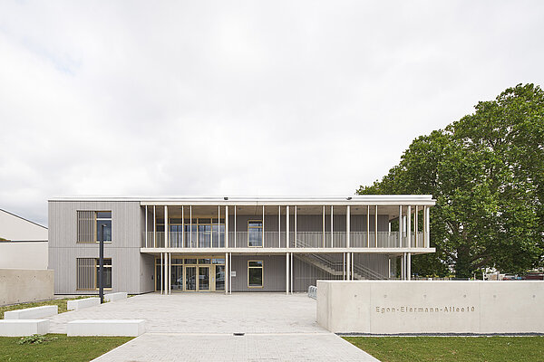 Das Bild zeigt die Südwestfassade mit heller Holzverkleidung, Balkon und dem Eingangsbereich mit Schriftzug.