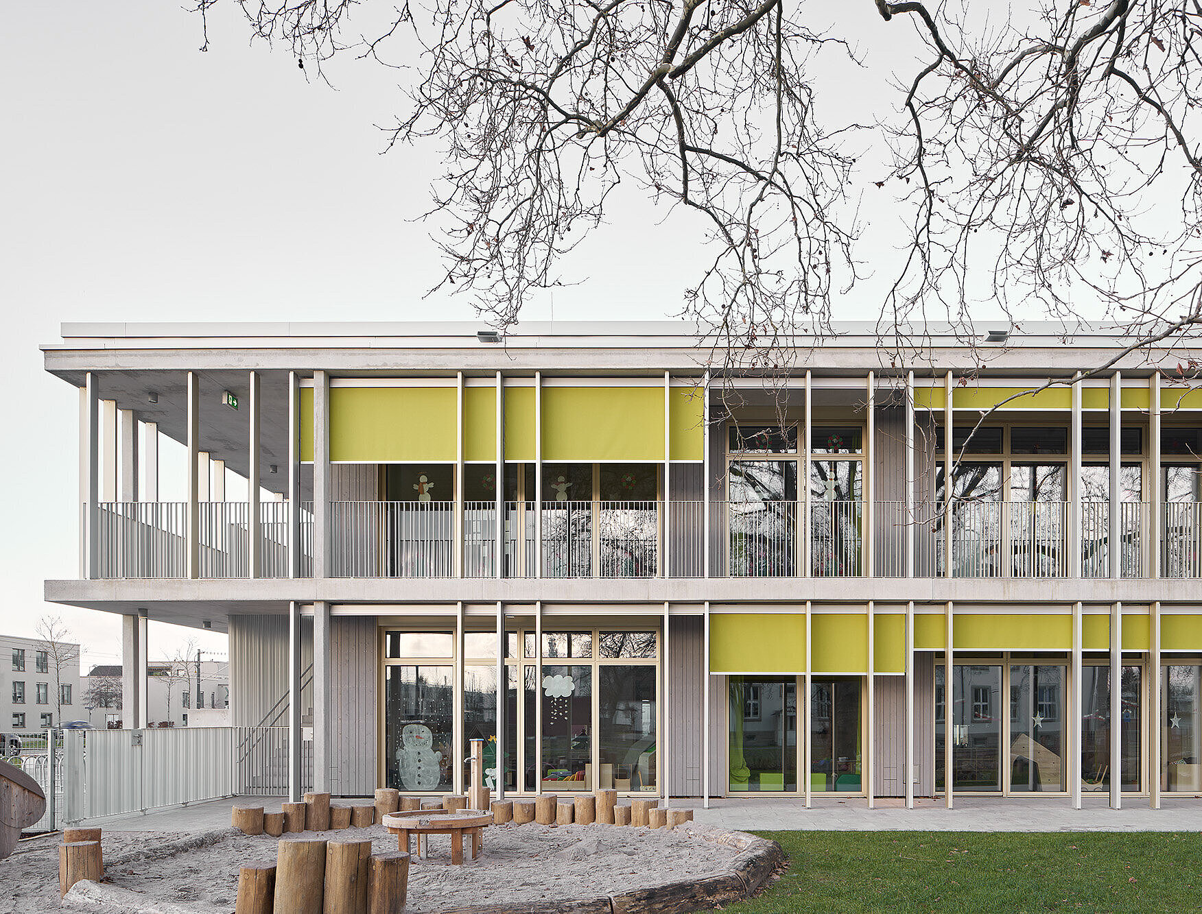 Das Foto zeigt einen Abschnitt der Südostfassade mit heller Holzverkleidung und auffälligem Sonnenschutz sowie einen Teil des Spielgartens.