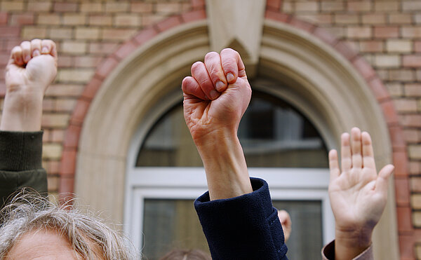 Drei Hände, zwei mit Faust, eine geöffnet als Symbol für das Hilfezeichen