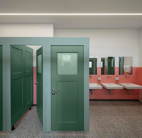 Blick auf zwei sanierte grüne Tailettentüren und drei Waschbecken mit Spiegel. 