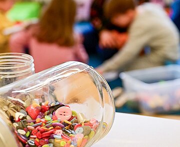 Im Vordergrund liegen Knöpfe in einem Glas, im Hintergrund in der Unschärfe spielen Kinder
