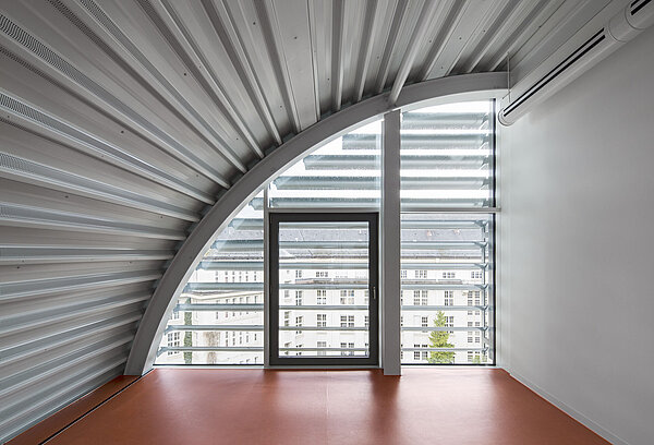 Zu sehen ist ein Teil des hellen, weißen Dachraums mit sichtbarer Stahlkonstruktion des Tonnendachs, verglasten Fronten und rotbraunem Linoleum-Boden.