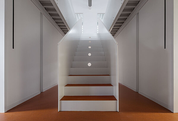 Blick auf den Treppenaufgang mit weißem Anstrich und rotbraunem Linoleum-Treppenbelag, der genau wie der restliche Bodenbelag gestaltet ist.