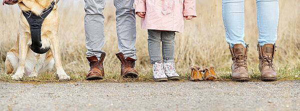 Straßenrand mit Wiese: Beine von einem Hund, zwei Erwachsenen und einem Kind und einem Paar Babyschuhe.